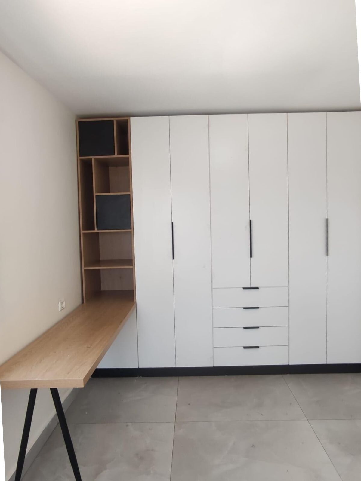 ארון קיר מעוצב בקווים מינימליסטיים, עם חזית לבנה וידיות שחורות, בשילוב שולחן עבודה מעץ ונישות פתוחות מעץ – פתרון מושלם המשלב אחסון, עבודה ועיצוב אחיד.