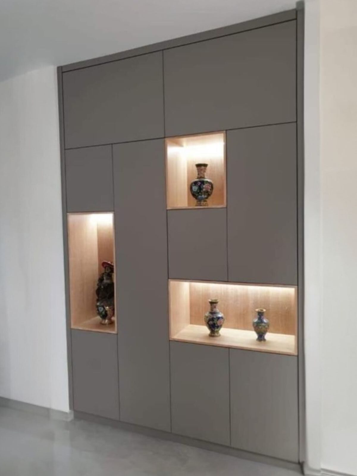 ארון קיר בעל חזית אחידה בגוון כהה, משולב נישות פתוחות בגימור עץ ותאורה פנימית, ליצירת חלוקה א־סימטרית ועומק חזותי.