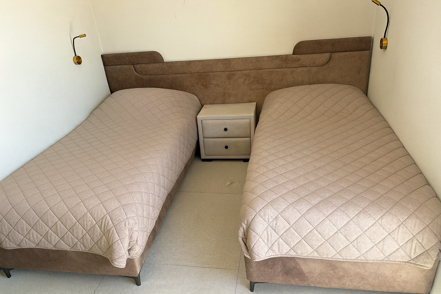 מיטות חדר שינה עם ראש מיטה מרופד בצע חום חמים בעיצוב רציף עם קו עליון מעוגל וקצוות מודגשים, בשילוב תאורת קיר אלגנטית