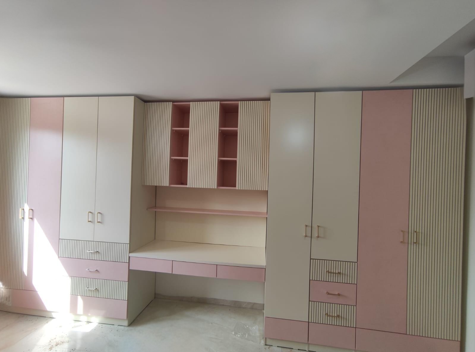 ארון בגדים בעיצוב נקי ומודרני בגוון בהיר, משולב עמדת עבודה צדדית עם משטח ומגירות בגוון ורוד רך. הארונות העליונים והנישות הפתוחות יוצרים פתרון אחסון חכם ופונקציונלי, תוך שמירה על מראה צעיר, מסודר ונעים לעין.