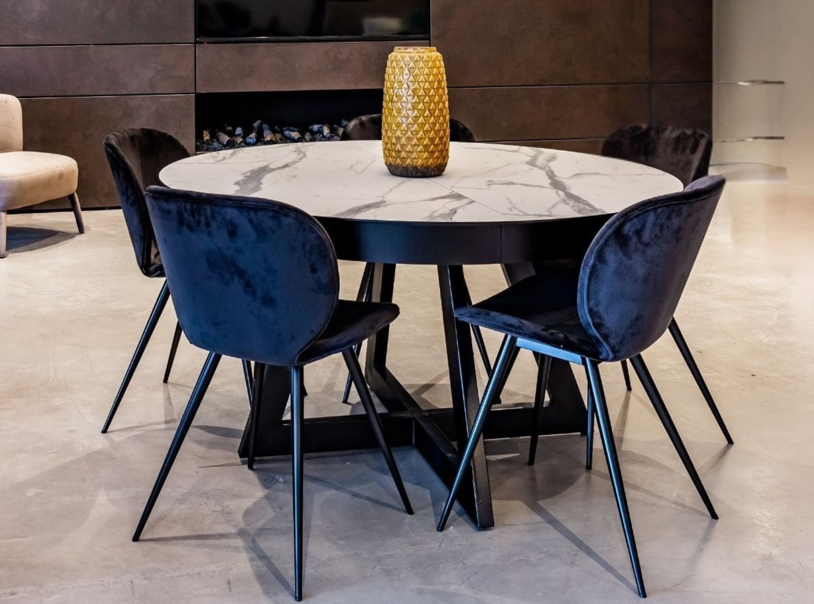 שולחן אוכל עגול עם משטח דמוי שיש ובסיס מתכת שחור, בשילוב כיסאות מרופדים כהים בעיצוב מעוגל.