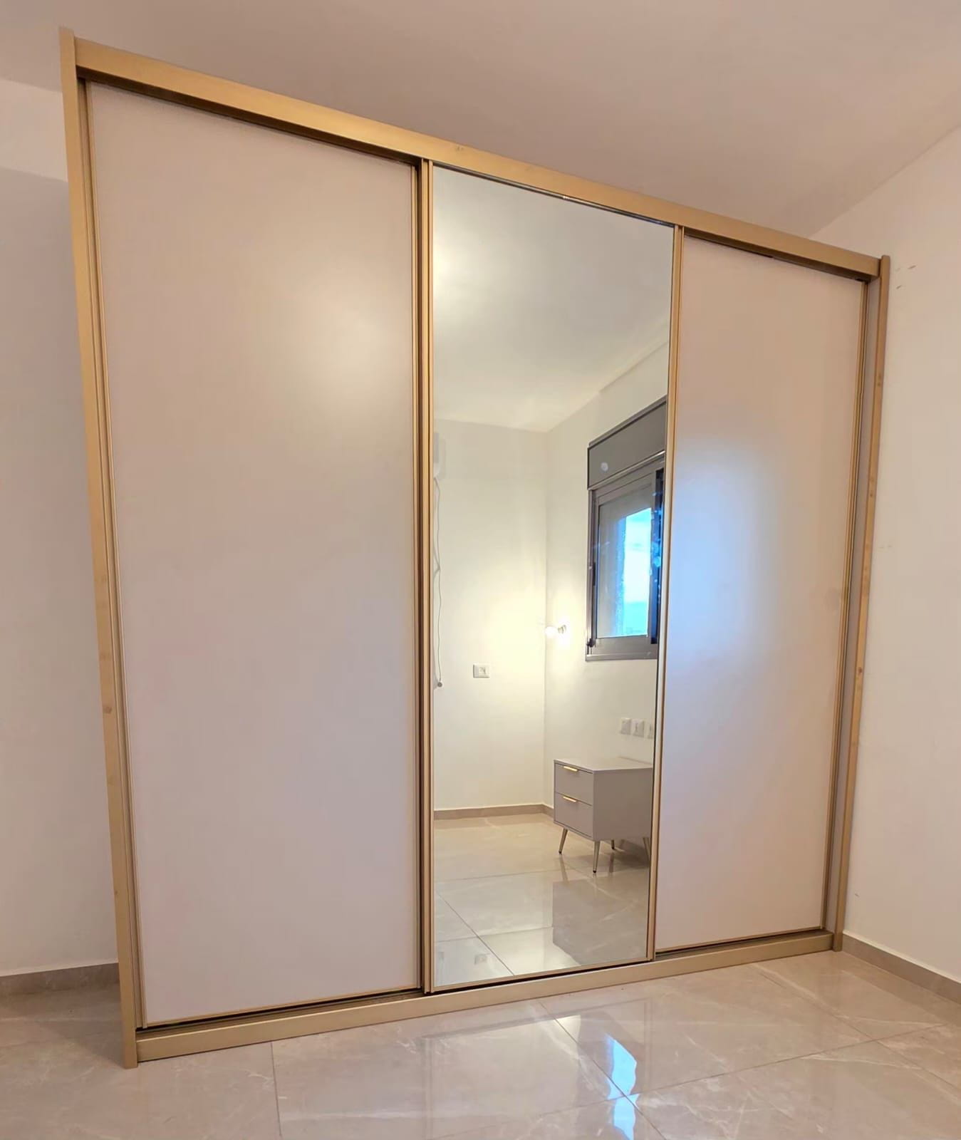 ארון הזזה רחב בעל שלוש דלתות בעיצוב מודרני ונקי. הארון משלב שתי דלתות צדדיות בגוון שמנת מט עם דלת מראה מרכזית גדולה המעניקה תחושת מרחב בחדר. כל דלתות הארון מוקפות במסגרת זהב עדינה ויוקרתית, המשתלבת עם המראה הכללי של החלל