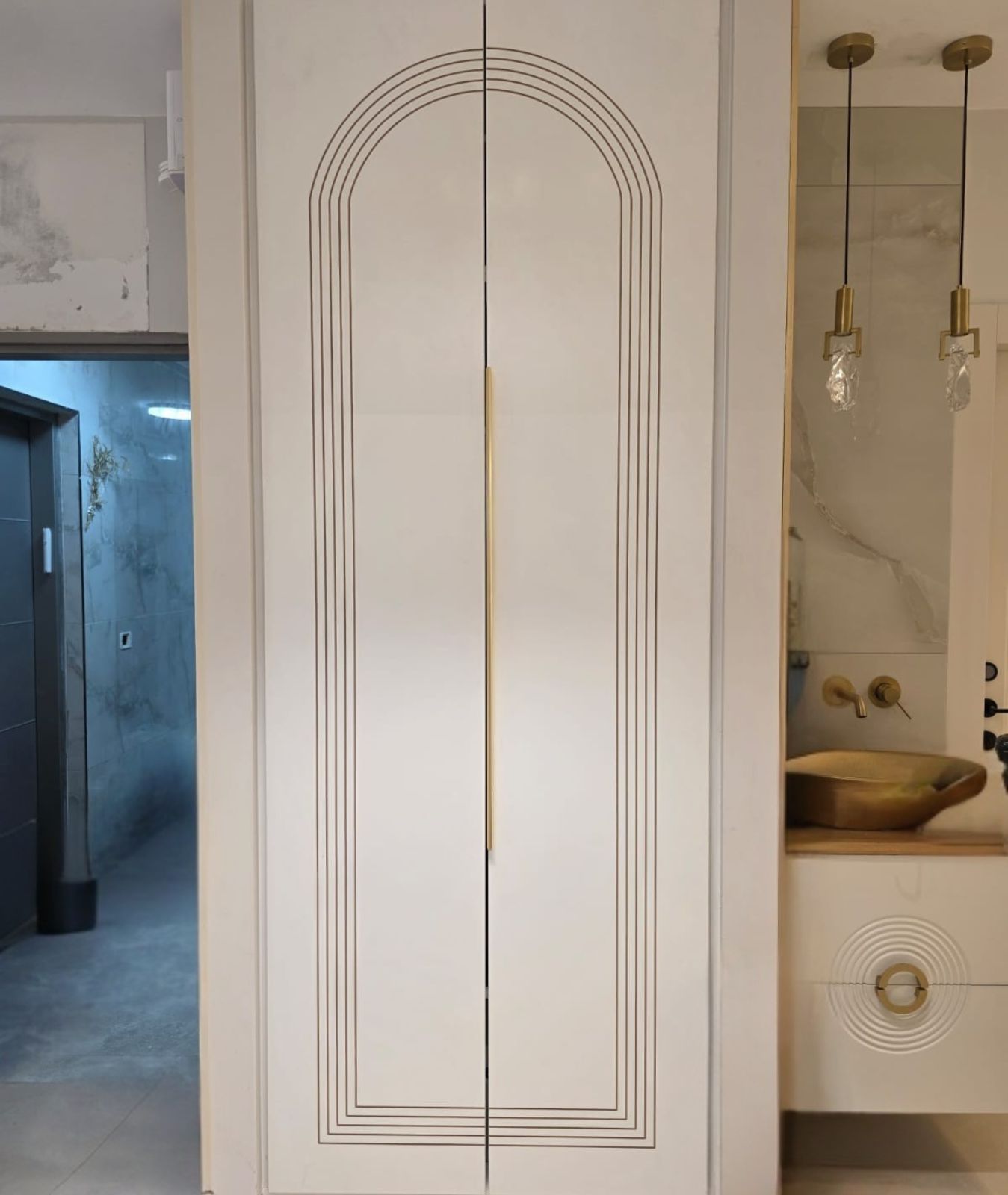 ארון יוקרתי בגוון שמנת מוקפד. דלתות הארון מעוטרות בחריטת פסים עדינה בצורת קשת גבוהה, המעניקה עומק ויזואלי ותחושת גובה. במרכז הדלתות מותקנת ידית זהב אנכית וארוכה המשתלבת עם הקו העיצובי היוקרתי. לצד הארון נראית יחידת כיור משולבת עם ברזי זהב ותאורה תואמת, היוצרים מראה הרמוני ומודרני בחלל.