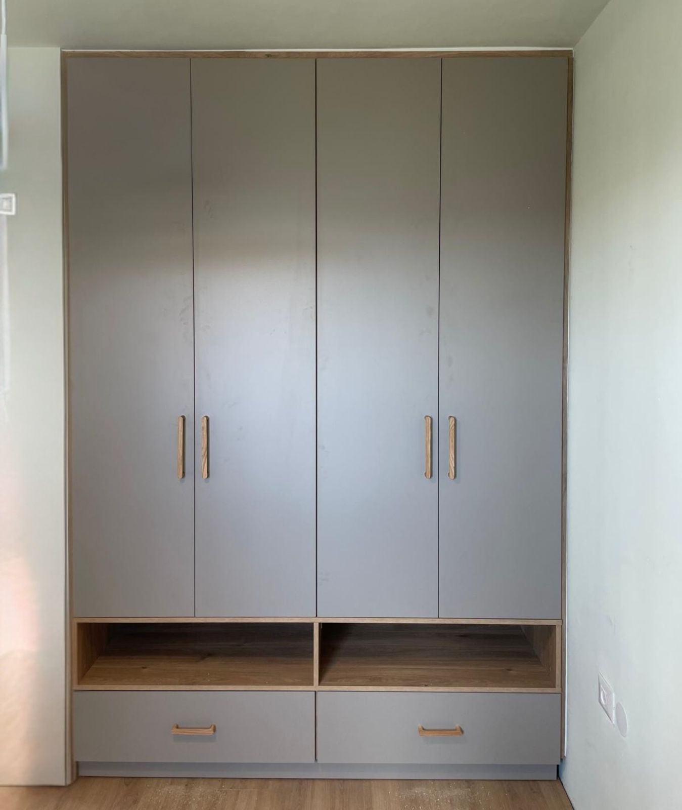 ארון קיר מעוצב ופונקציונלי בגוון גרייז' מודרני, המשלב פתרונות אחסון מגוונים. הארון כולל ארבע דלתות גבוהות עם ידיות עץ אנכיות, ומתחתן שתי נישות מדפים פתוחות מעץ בגוון טבעי וחמים. בחלקו התחתון משולבות שתי מגירות רחבות עם ידיות עץ תואמות, למראה נקי, מאורגן וניצול חכם של גובה החדר.