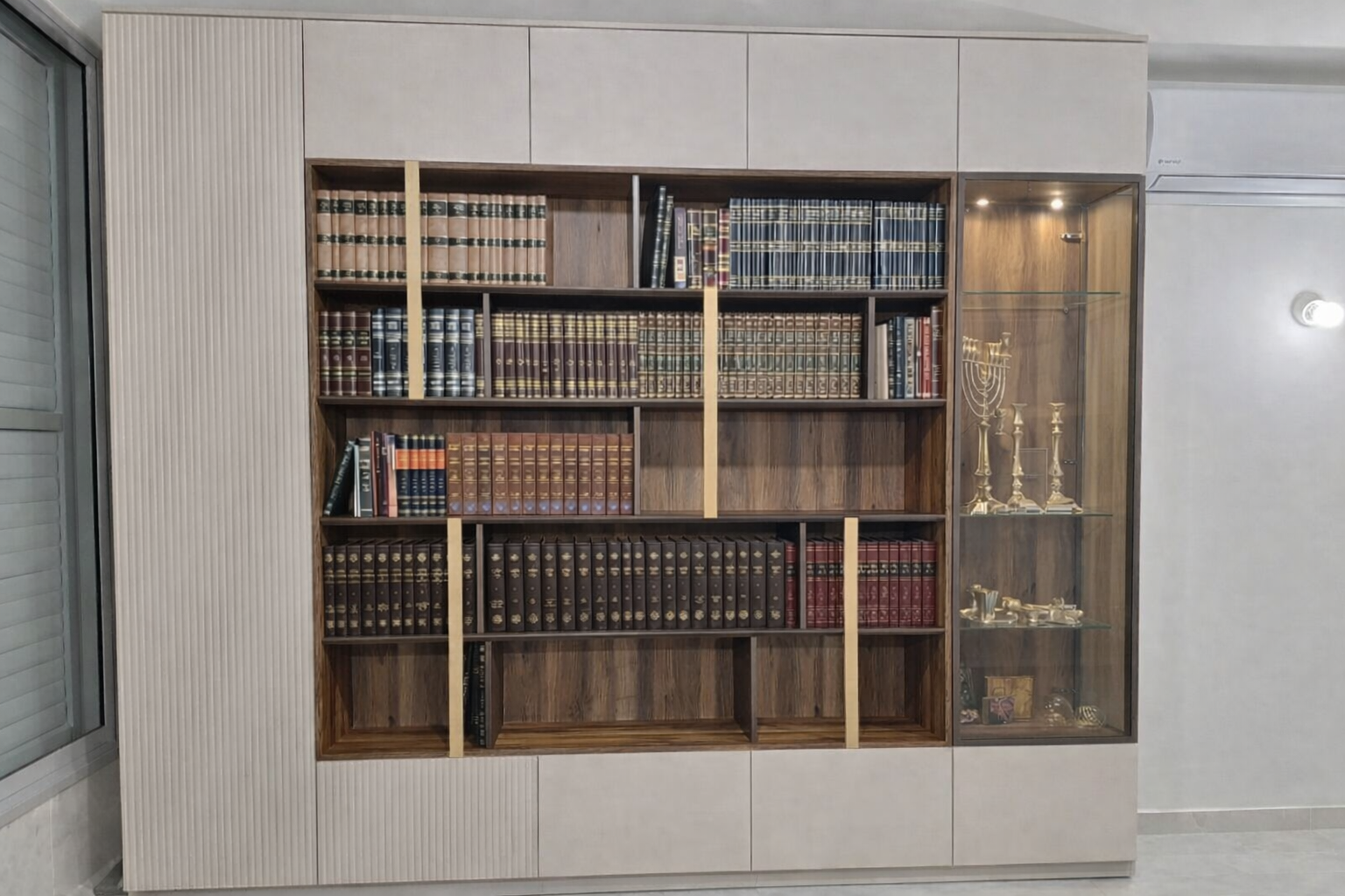 ספריית קודש מעוצבת המשלבת נראות יוקרתית עם פתרונות אחסון פרקטיים. עם מדפים עץ אלגנטי במרכז המעוטרים בפסי הפרדה מוזהבים ואנכיים המעניקים מראה יוקרתי, המוקפים במערכת ארונות אחסון סגורים בגימור בהיר עם טקסטורת פסים אנכיים (חריטות לייזר). בעוד הויטרינה המשולבת בצד מוסיפה אלמנט תצוגה מואר.