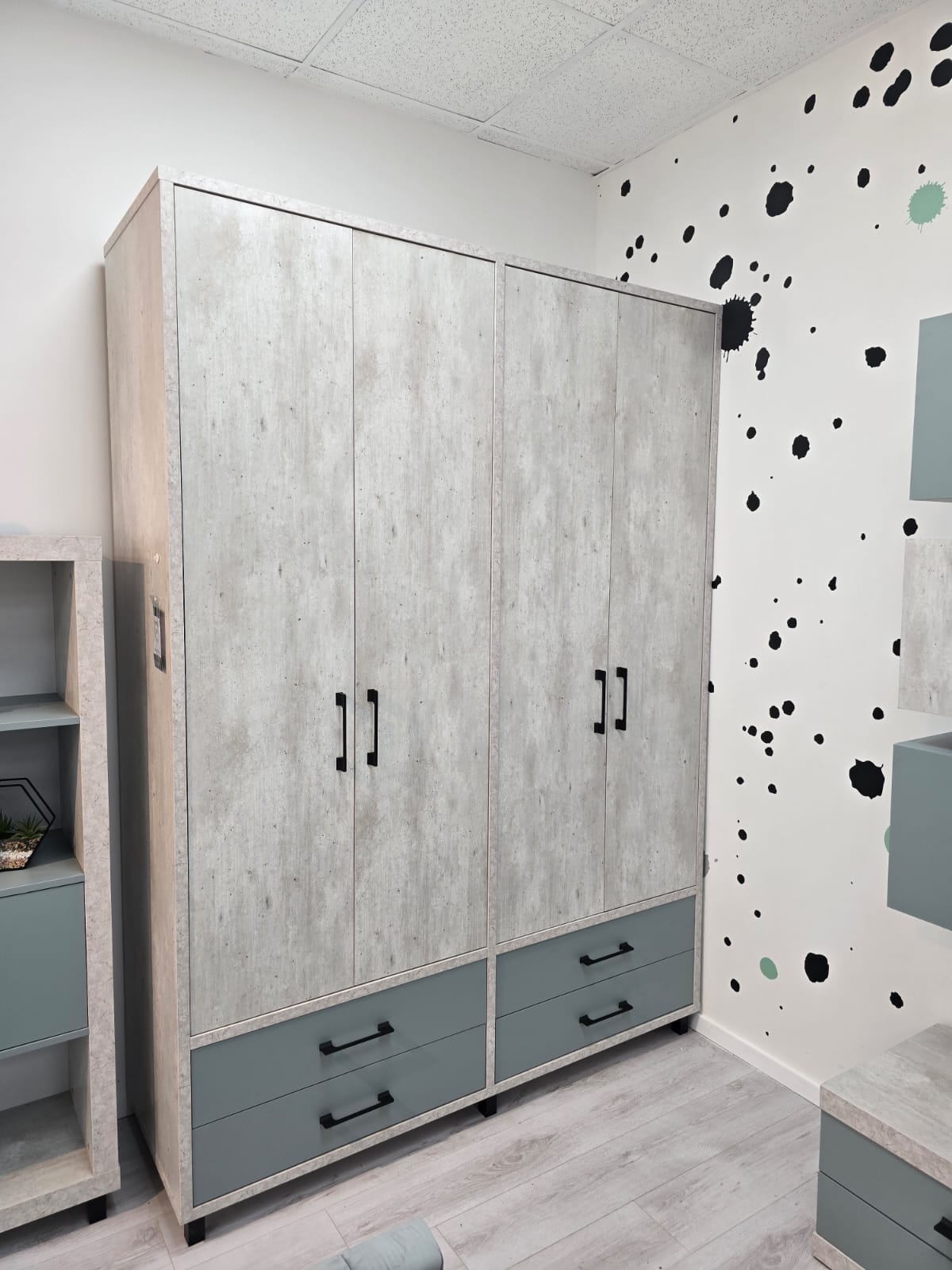 ארון בגדים בגימור עץ בהיר עם ארבע דלתות וידיות שחורות, ובחלק התחתון שתי מגירות בצבע אפור-ירקרק