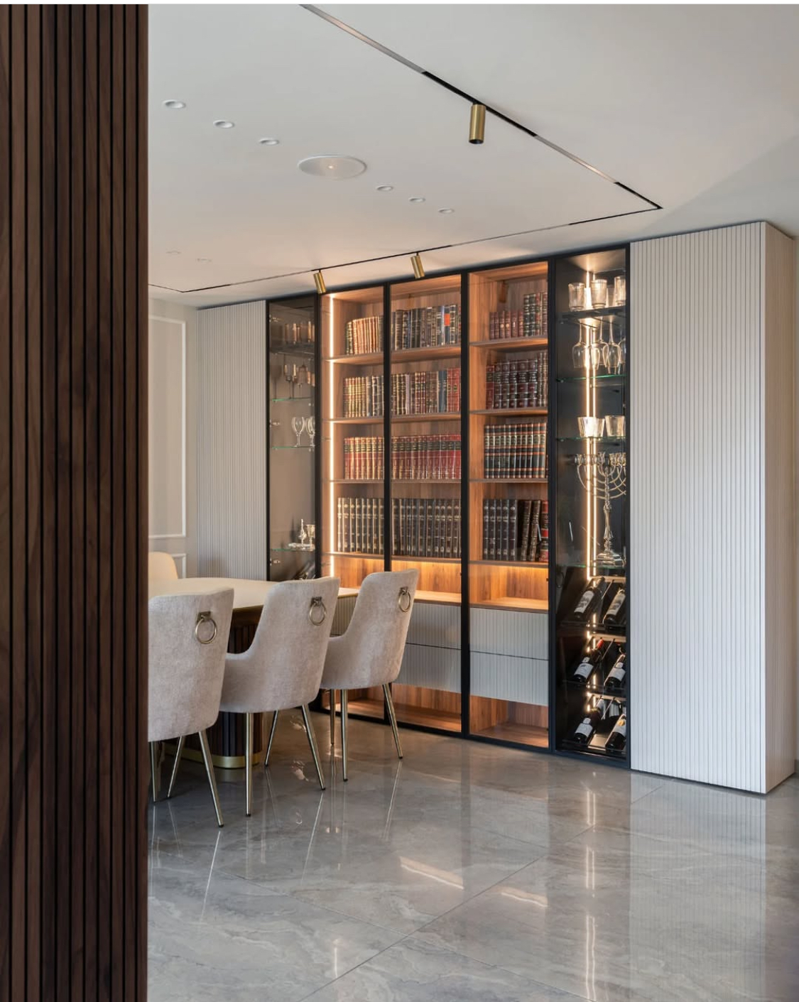 ספריית קודש יוקרתית המשלבת פתרונות תצוגה ואחסון רחבים במראה מודרני ונקי. היחידה כוללת מדפי עץ במרכז עם תאורה פנימית חמימה, המוקפת בוויטרינות זכוכית גבוהות עם מסגרת שחורה דקה להצגת כלי יודאיקה ובקבוקי יין. צידי הספרייה כוללים ארונות אחסון סגורים בגימור בהיר עם טקסטורה מחורצת (סטריפים), המעניקים מראה אלגנטי וניצול מקסימלי של חלל הקיר.