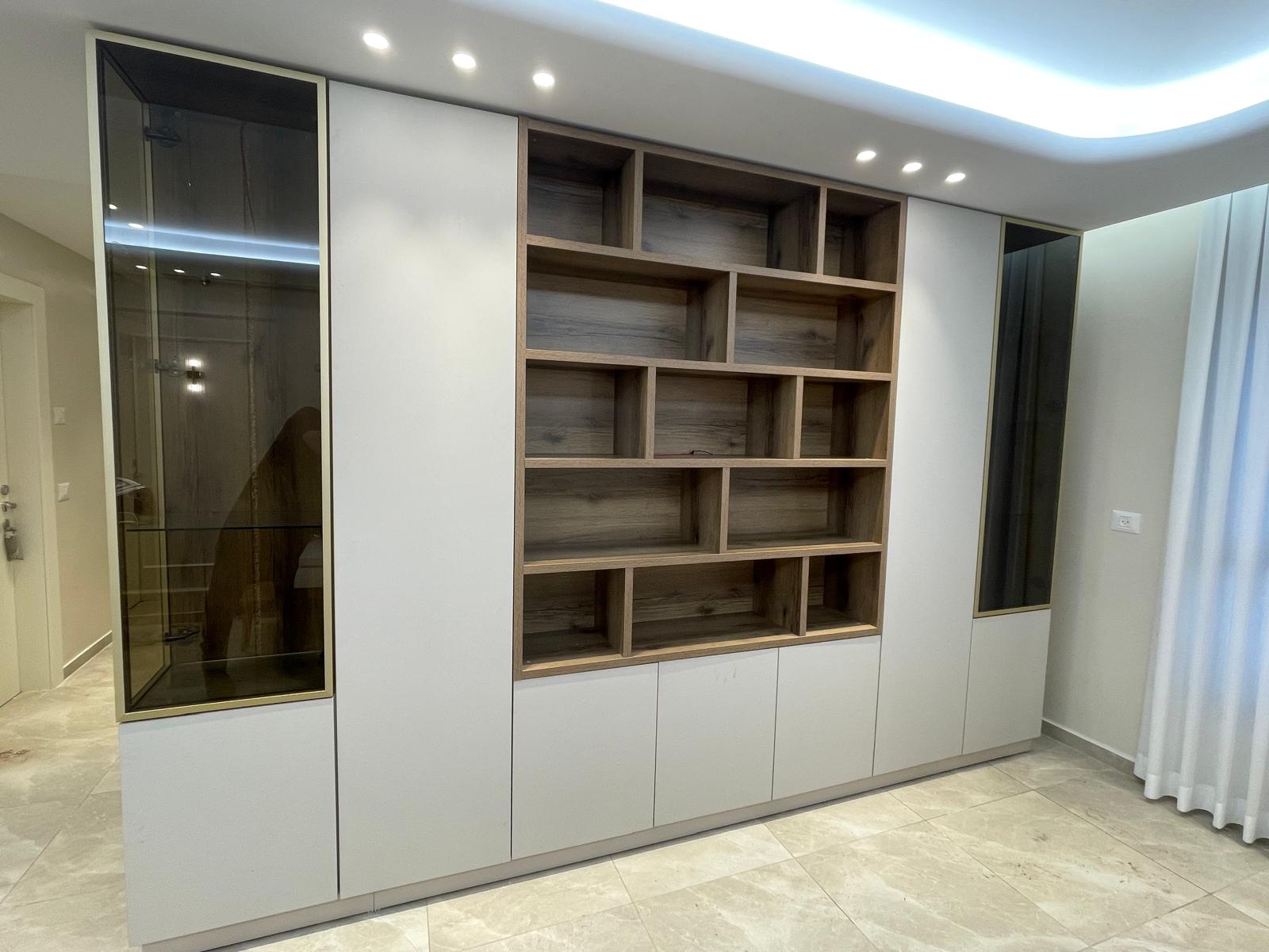 ספריית קודש מודרנית ויוקרתית המשלבת פתרונות תצוגה ואחסון רחבים. היחידה כוללת כוורת מדפים פתוחה מעץ בגוון חמימים במרכז, המוקפת בארונות אחסון סגורים בגימור לבן מט נקי (פתיחת טאץ'). בשני צידי הספרייה משולבות ויטרינות זכוכית גבוהות עם מסגרת זהב דקה ותאורה פנימית, המעניקות מראה אלגנטי וניצול מקסימלי של חלל הקיר.