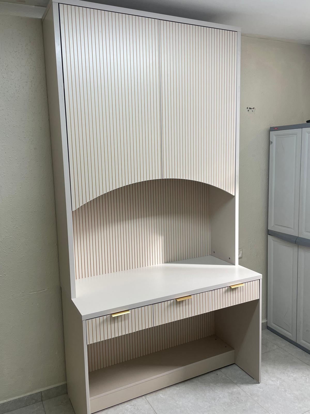 שולחן כתיבה עם שילוב ארון אחסון עליון. היחידה כוללת שלוש מגירות תחתונות עם ידיות זהב אלגנטיות וחזיתות מחורצות. ארון האחסון העליון מעוטר בחזית פסים תואמת עם סיומת קשתית מעל משטח העבודה, המעניקה מראה מודרני, נקי וניצול חכם של שטח הקיר.