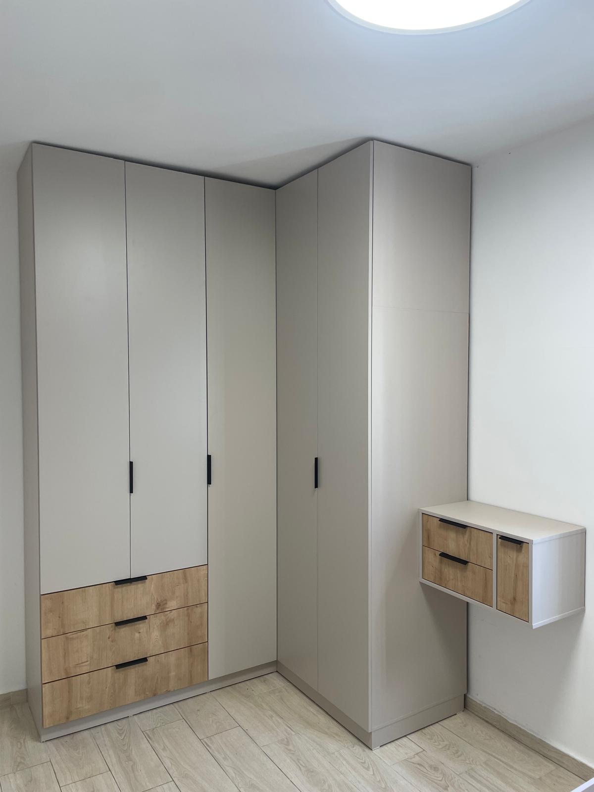 ארון בגדים פינתי ומעוצב בגוון גרייז' נקי עם ידיות שחורות מינימליסטיות, ומשלב שלוש מגירות חיצוניות בחזית עץ טבעי למראה חמים ומודרני. בצדו מחוברת יחידת מגירות "מרחפת" תואמת עם חזיתות עץ, המעניקה פתרון אחסון נוסף וקליל מבלי להעמיס על רצפת החדר.