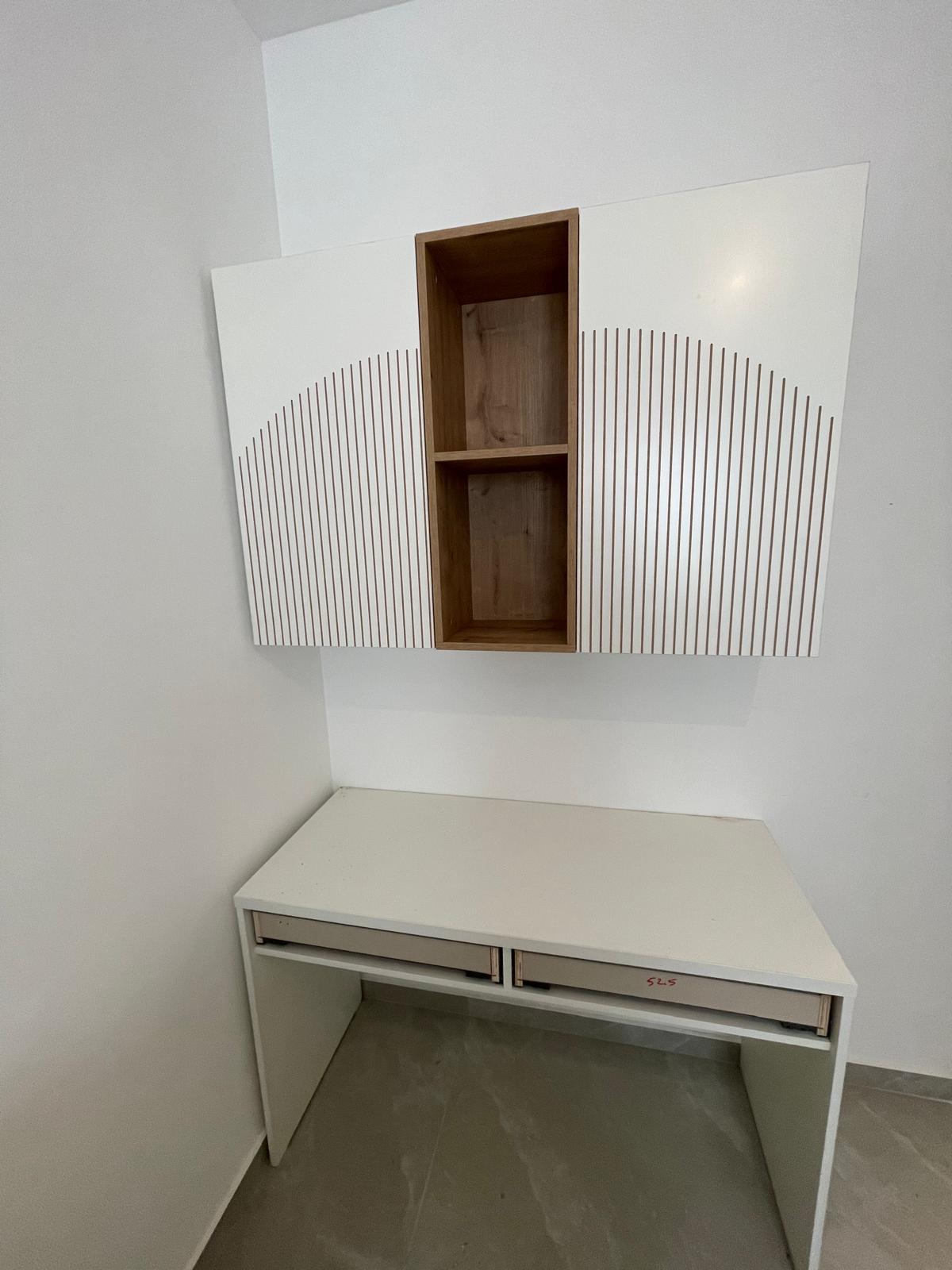 שולחן כתיבה בהיר עם מגירות אחסון דקות, ומעליו כוורת תלויה. היחידה משלבת מדפי עץ פתוחים במרכז לצד ארונות אחסון סגורים בגימור לבן, המעוטרים בדוגמת חצי עיגול של חירוץ עיצובי.