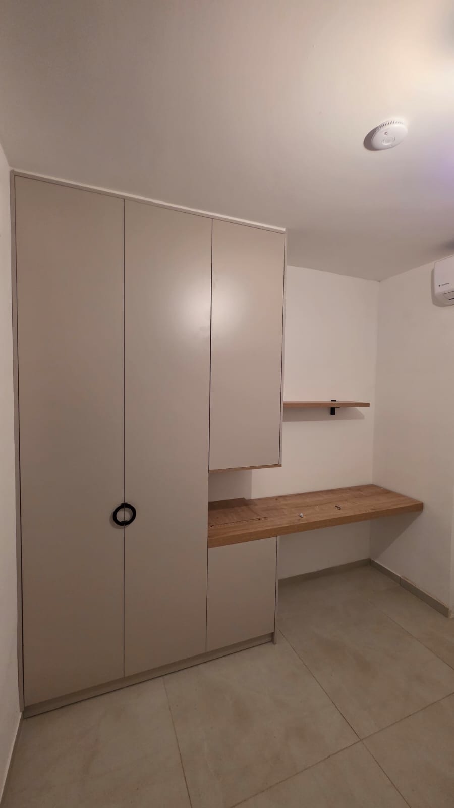 ארון בגדים גבוה בגוון גרייז' בהיר עם ידית עגולה שחורה, המחובר ליחידת שולחן כתיבה "מרחף" מעץ טבעי. העיצוב כולל ארונות אחסון עליונים ותחתונים סגורים המשתלבים עם פינת העבודה, למראה מודרני, נקי ופרקטי