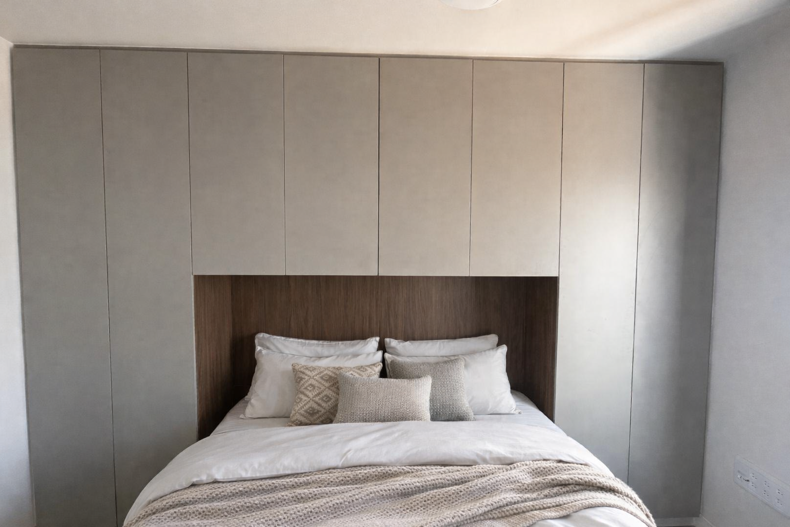ערכת ארונות קיר רחבה בגוון גרייז' העוטפת מיטה זוגית ומנצלת את כל שטח הקיר לאחסון מקסימלי. המיטה משולבת בתוך נישה פנימית מחופה עץ חמים, המשלבת בין פרקטיקה גבוהה למראה מודרני ונקי.